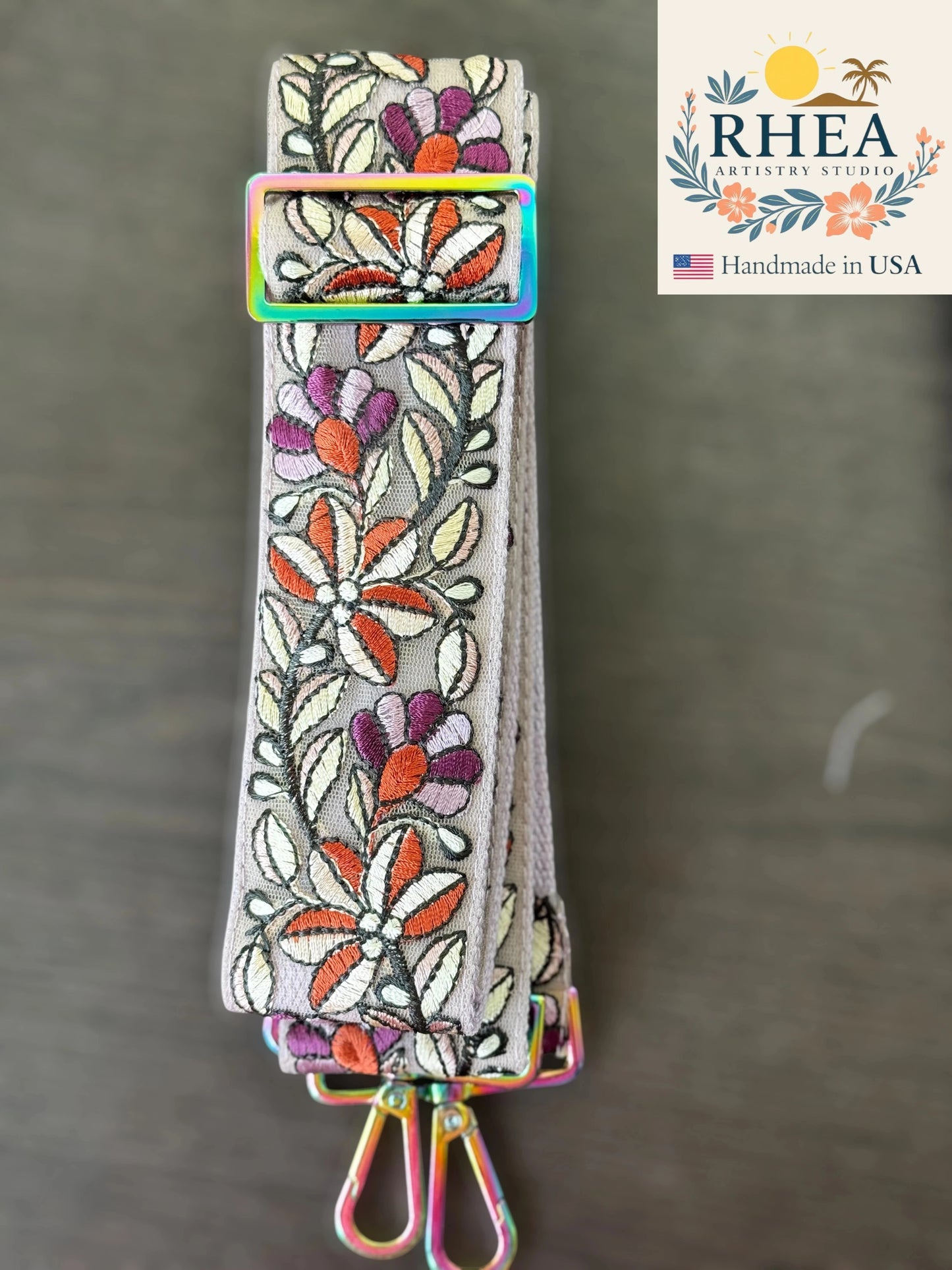 Embroidered purse strap with floral patterns on a gray background, featuring the RHEA brand.