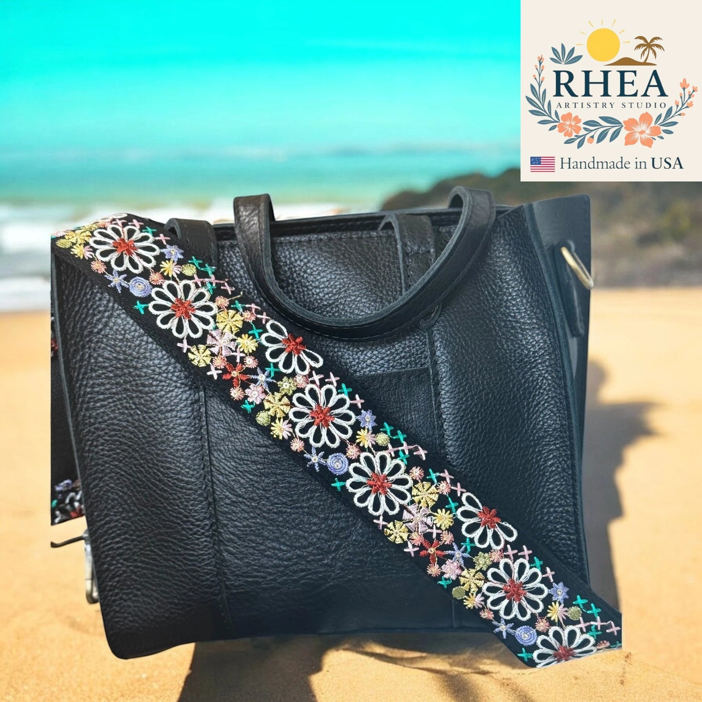 Black handbag with a colorful floral strap on a beach background, featuring the RHEA Artistry Studio logo.