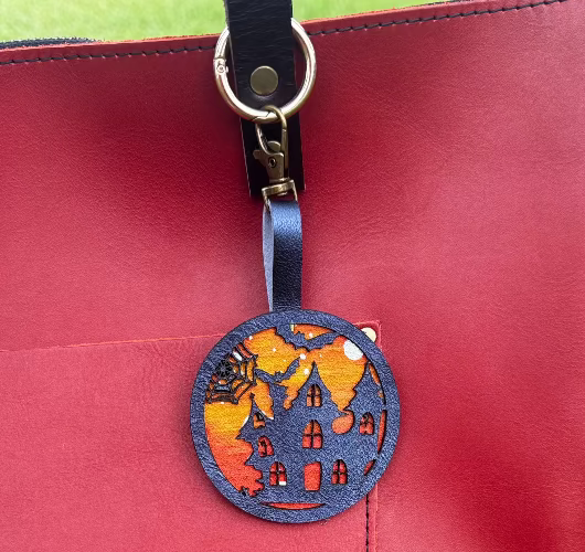 A handpainted leather charm with a halloweendesign, attached to a red leather bag.