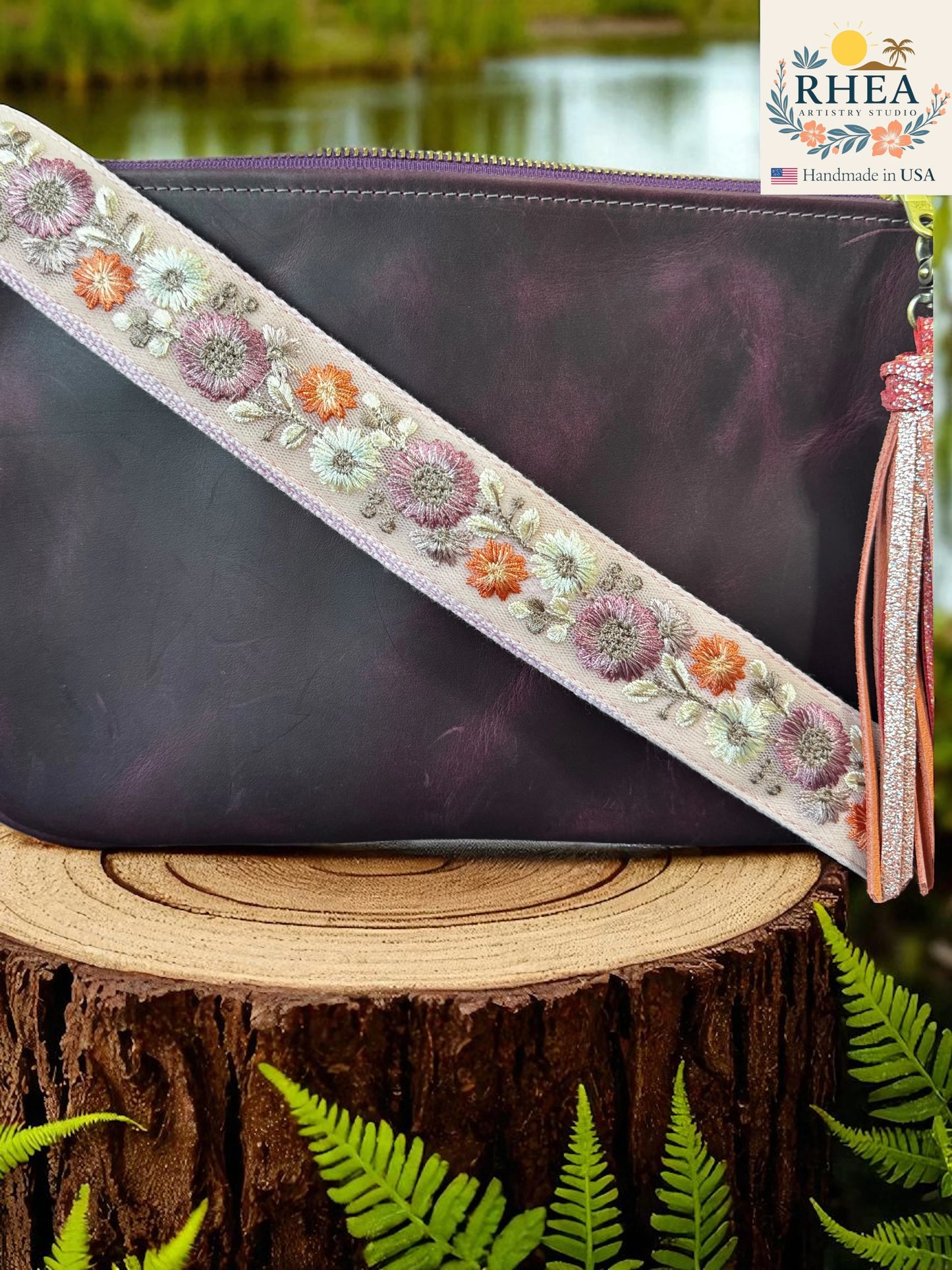 Purple clutch bag with floral strap on a wooden stump with ferns and blurred water background