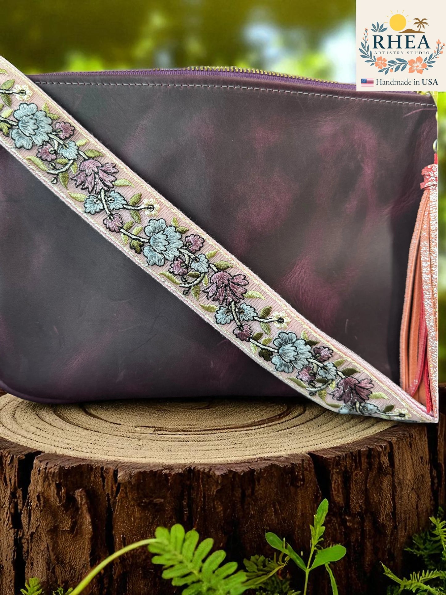 Purple pouch with floral leather bag strap on a wooden surface, RHEA brand visible