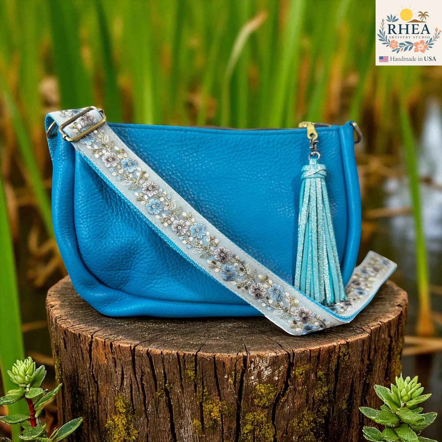 Blue handbag with decorative artisan strap and tassel on a wooden stump with greenery in the background, featuring the RHEA brand
