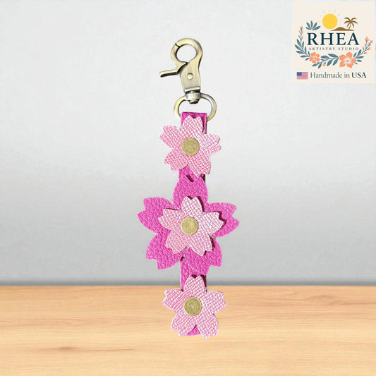 Pink flower-shaped leather purse charm with a clip on a wooden surface, branded 'RHEA'.