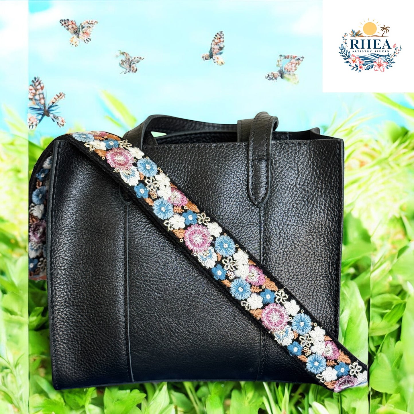 A black leather tote with an embroidered strap featuring blue, pink, and white flowers on a black background, against a bright grassy backdrop with butterflies in the sky.