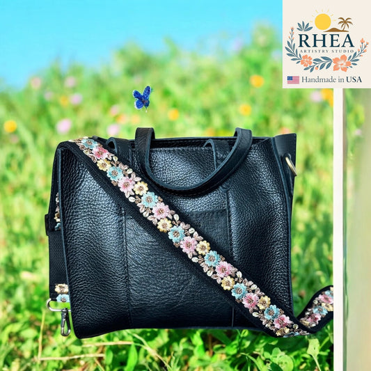 Black leather handbag with floral strap in a grassy field, featuring the RHEA brand.