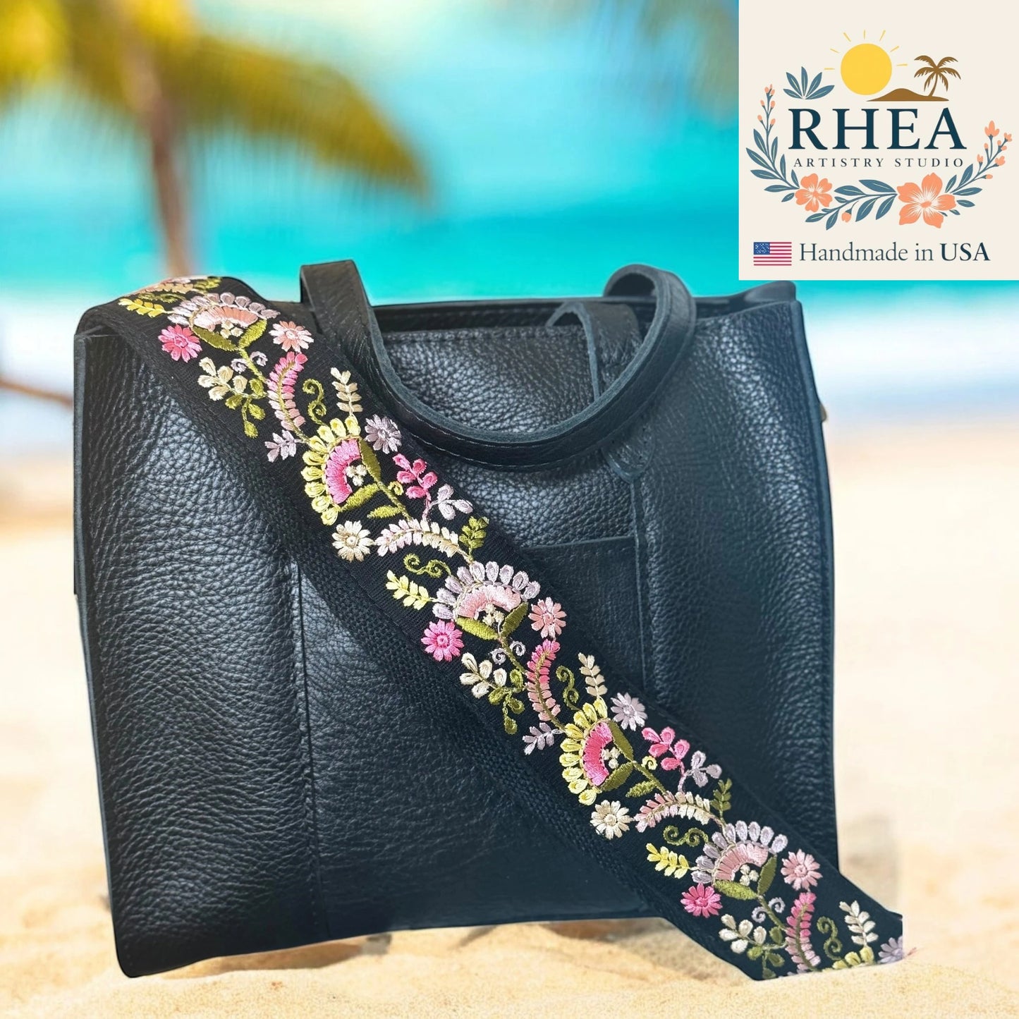 Black leather bag with floral embroidery strap on a beach background, featuring RHEA Artistry Studio logo.