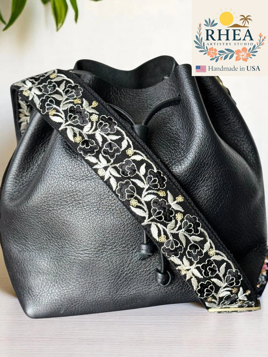Black leather bag with floral embroidery leather purse strap on a white surface, RHEA brand visible.