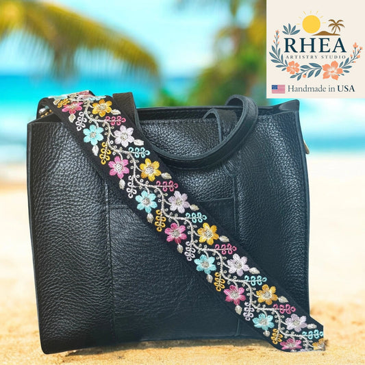 Black leather handbag with a colorful floral bag strap on a beach background, featuring the RHEA Artistry Studio logo.