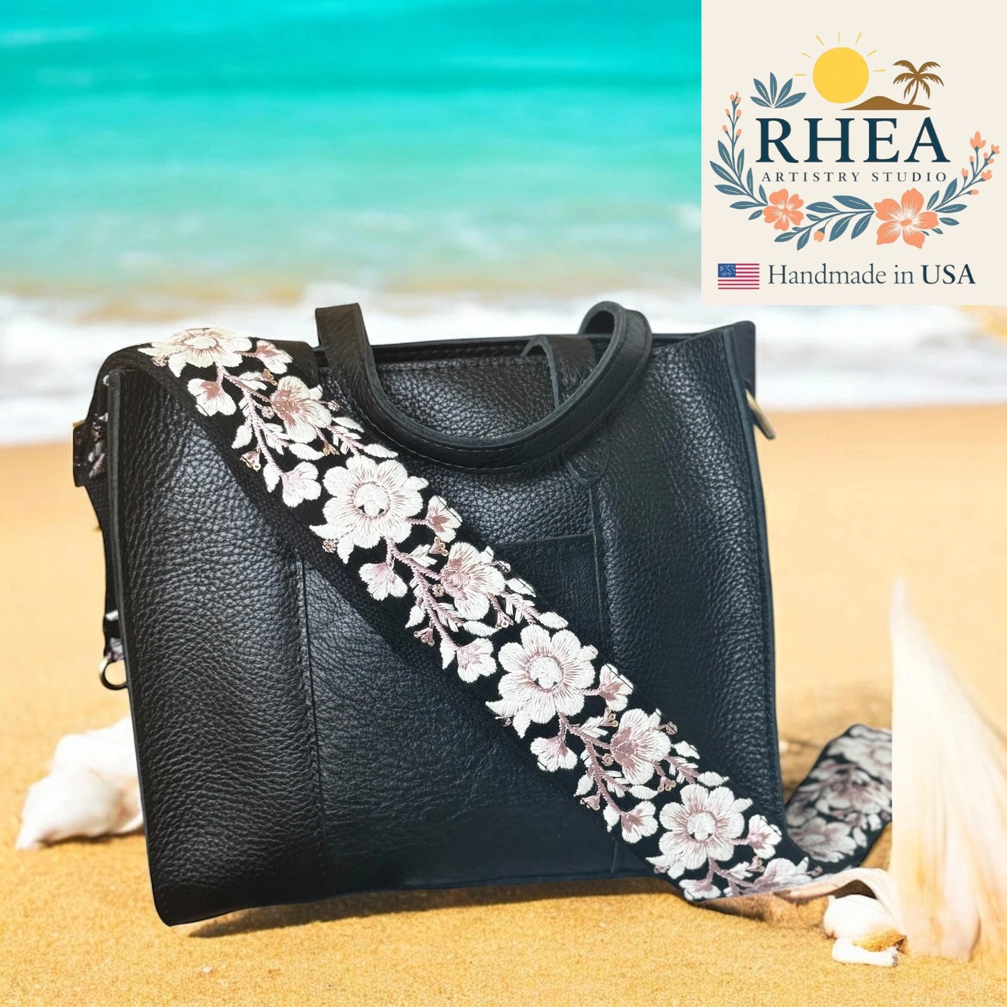 Black handbag with floral strap on a beach, featuring RHEA Artistry Studio logo.