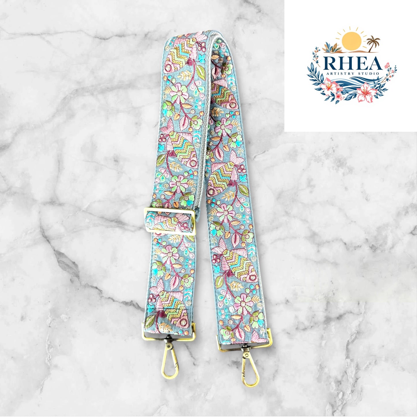An adjustable embroidered purse strap with floral patterns and gold-colored hardware, displayed on a gray background.