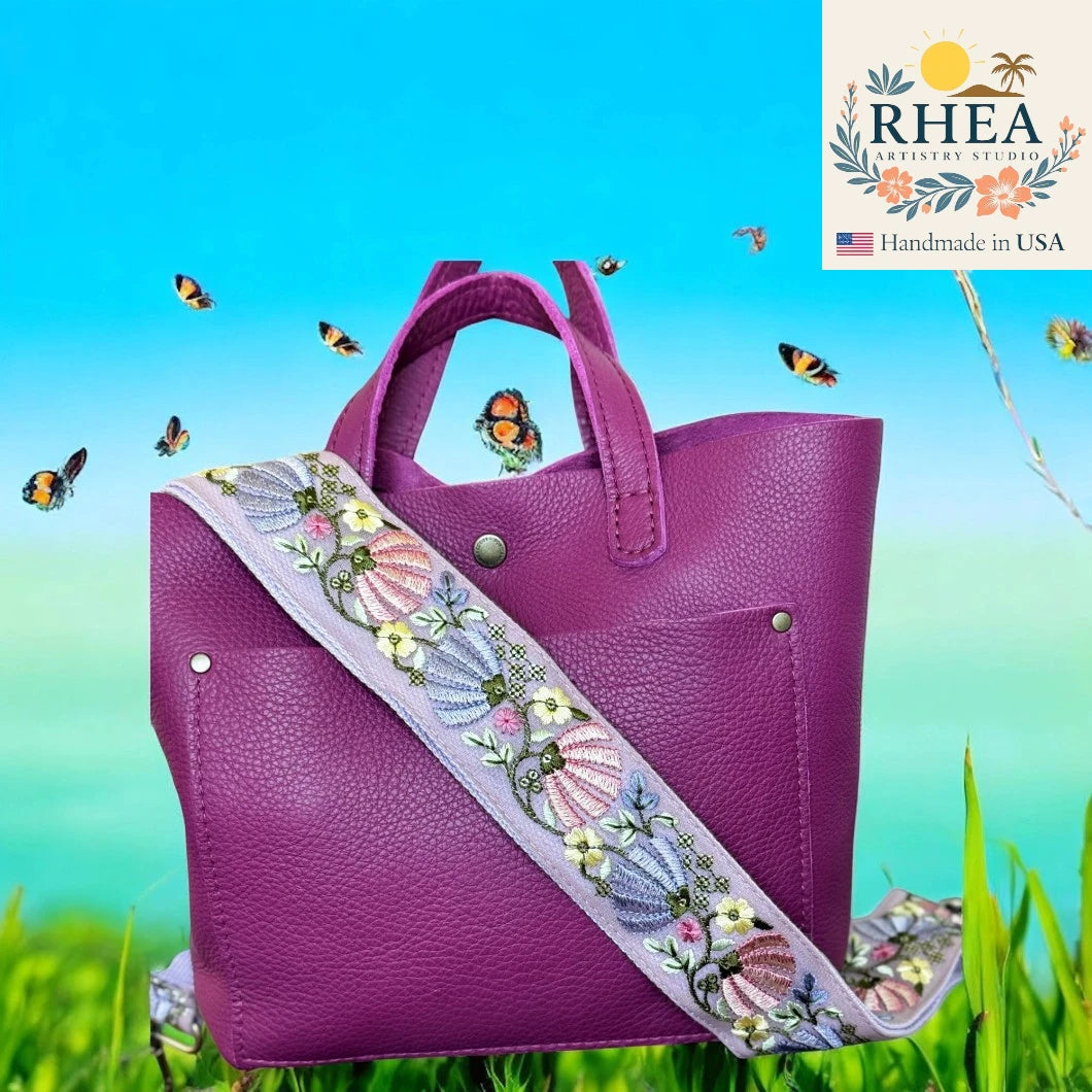 Purple handbag with an embroidered strap on a grassy background, featuring the RHEA brand.