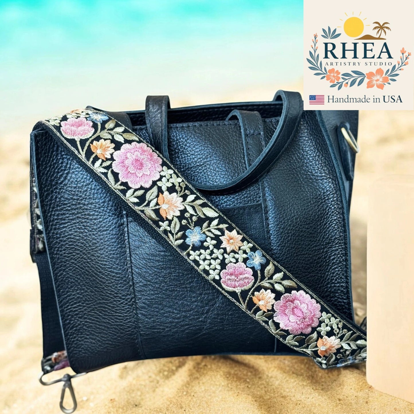 Black leather handbag with floral purse strap on a beach background, featuring the RHEA Artistry Studio logo.