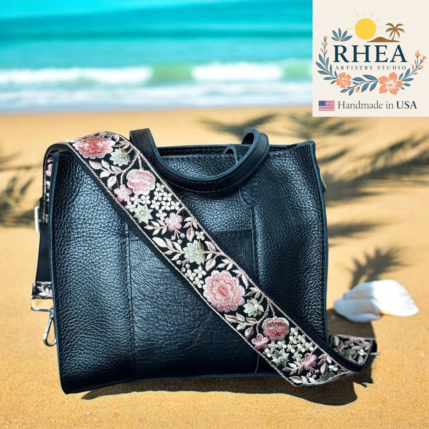 Black leather handbag with floral purse strap on a sandy beach, featuring RHEA Artistry Studio logo.