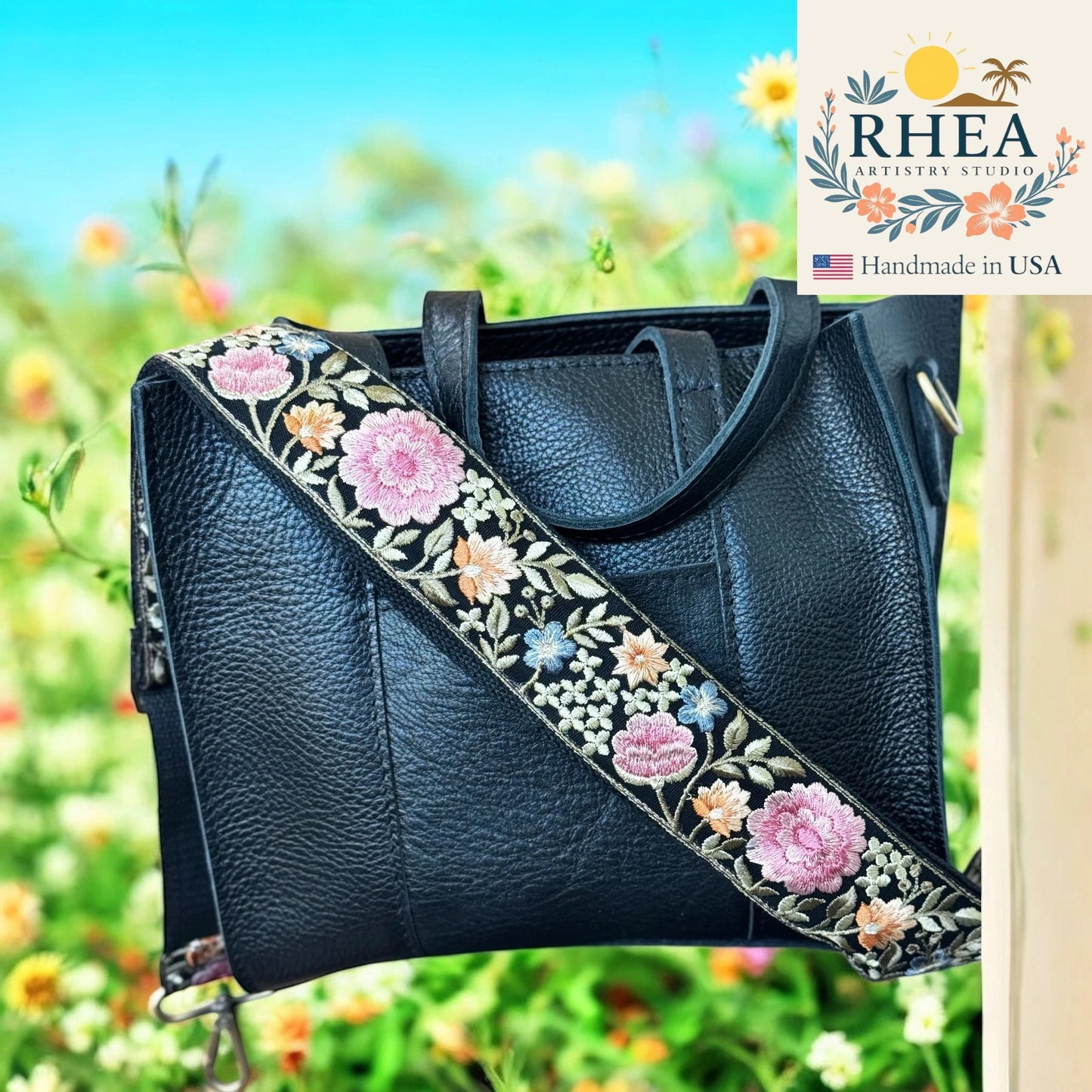 Black leather handbag with floral purse embroidery strap against a blurred natural background, featuring the RHEA Artistry Studio logo.