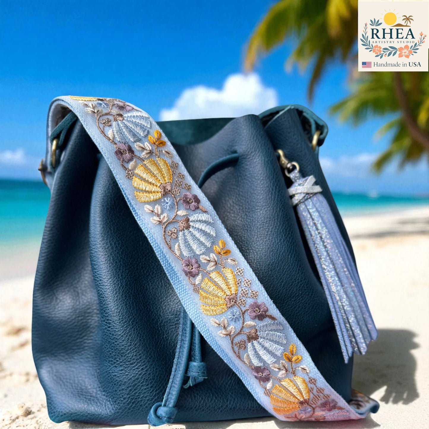 Blue handbag with an embroidered bag strap on a beach, featuring the RHEEA brand.