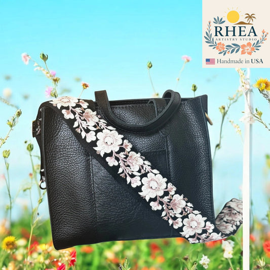 Black handbag with floral strap in a field of flowers, featuring the RHEA brand.