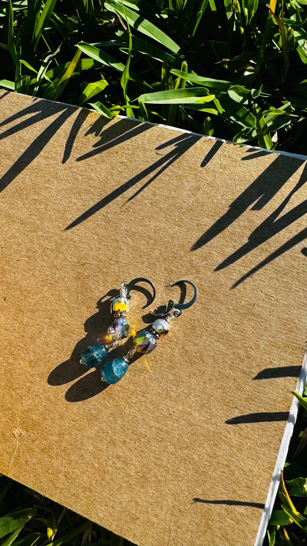 A pair of silver drop earrings with blue and multicolored gemstones, displayed on a brown card placed on grass.