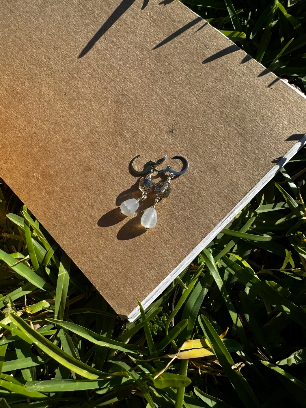A pair of pale blue silver drop earrings with a cathedral-like design, displayed on a brown paper surface, with blurred greenery in the background.