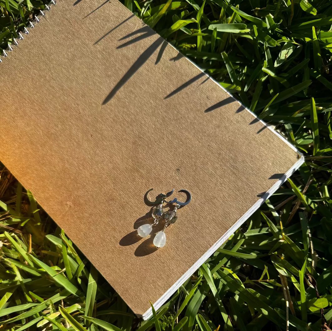 A pair of pale blue silver drop earrings with a cathedral-like design, displayed on a brown paper surface, with blurred greenery in the background.
