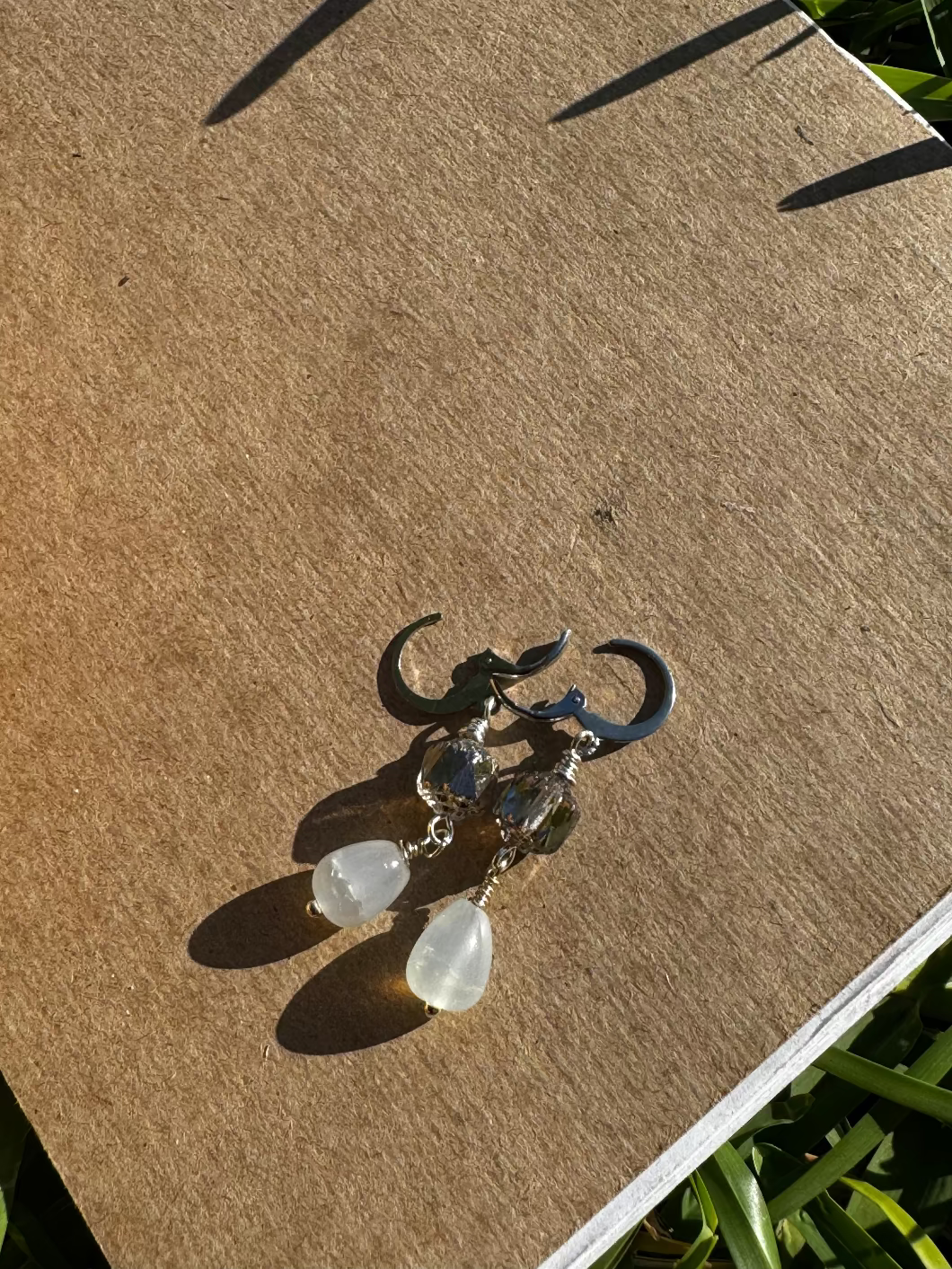 A pair of pale blue silver drop earrings with a cathedral-like design, displayed on a brown paper surface, with blurred greenery in the background.