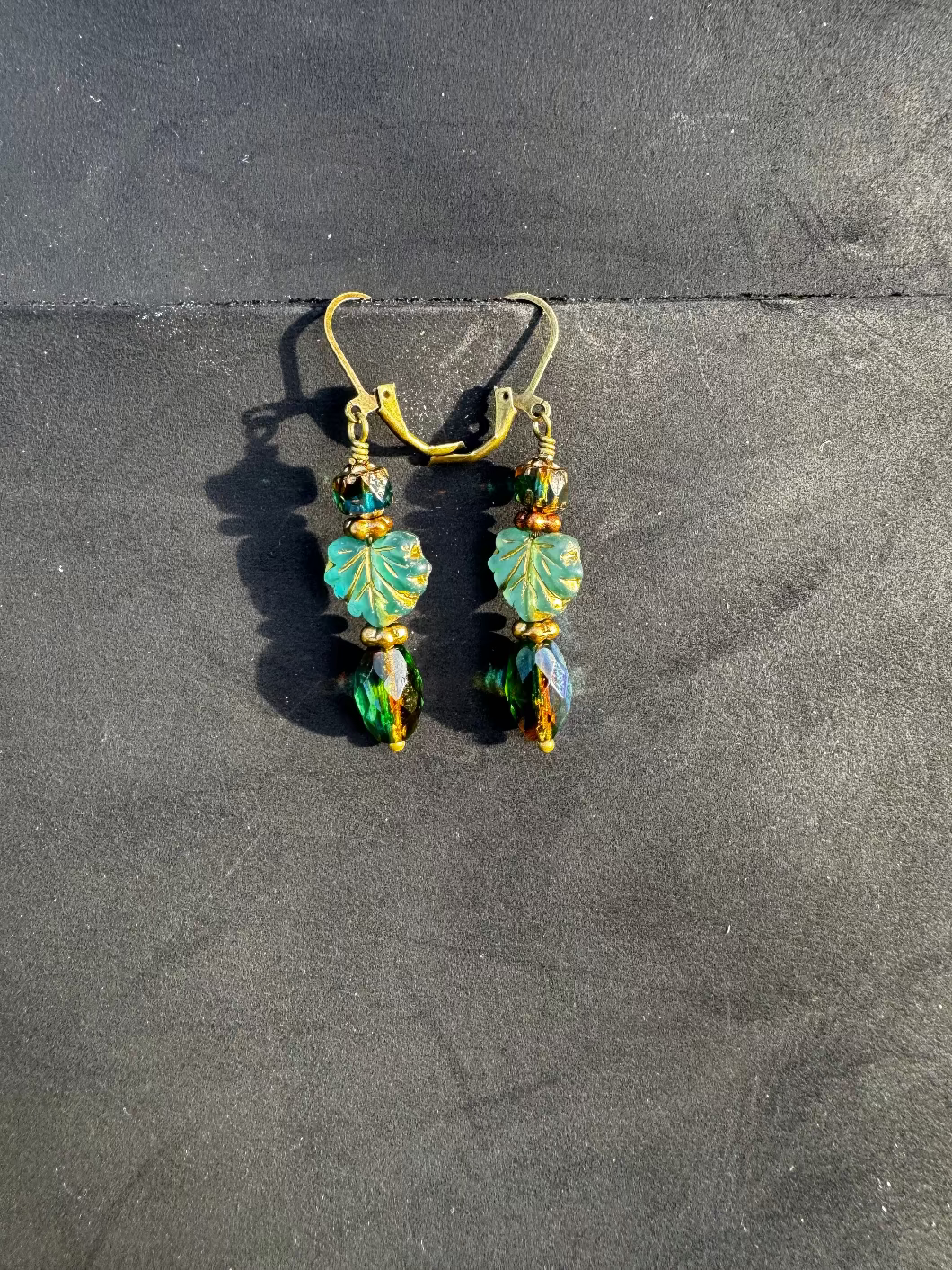 A pair of handcrafted earrings with green and blue czech glass beads, antique bronze finish, and brass hardware on a textured background.