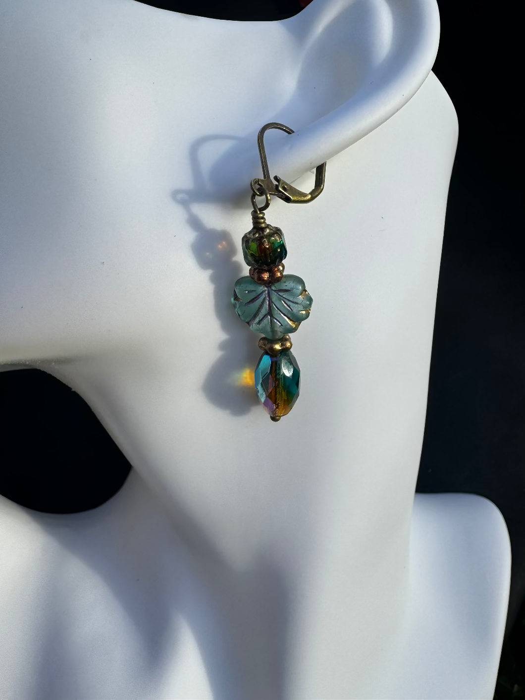 A pair of handcrafted earrings with green and blue czech glass beads, antique bronze finish, and brass hardware on a textured background.