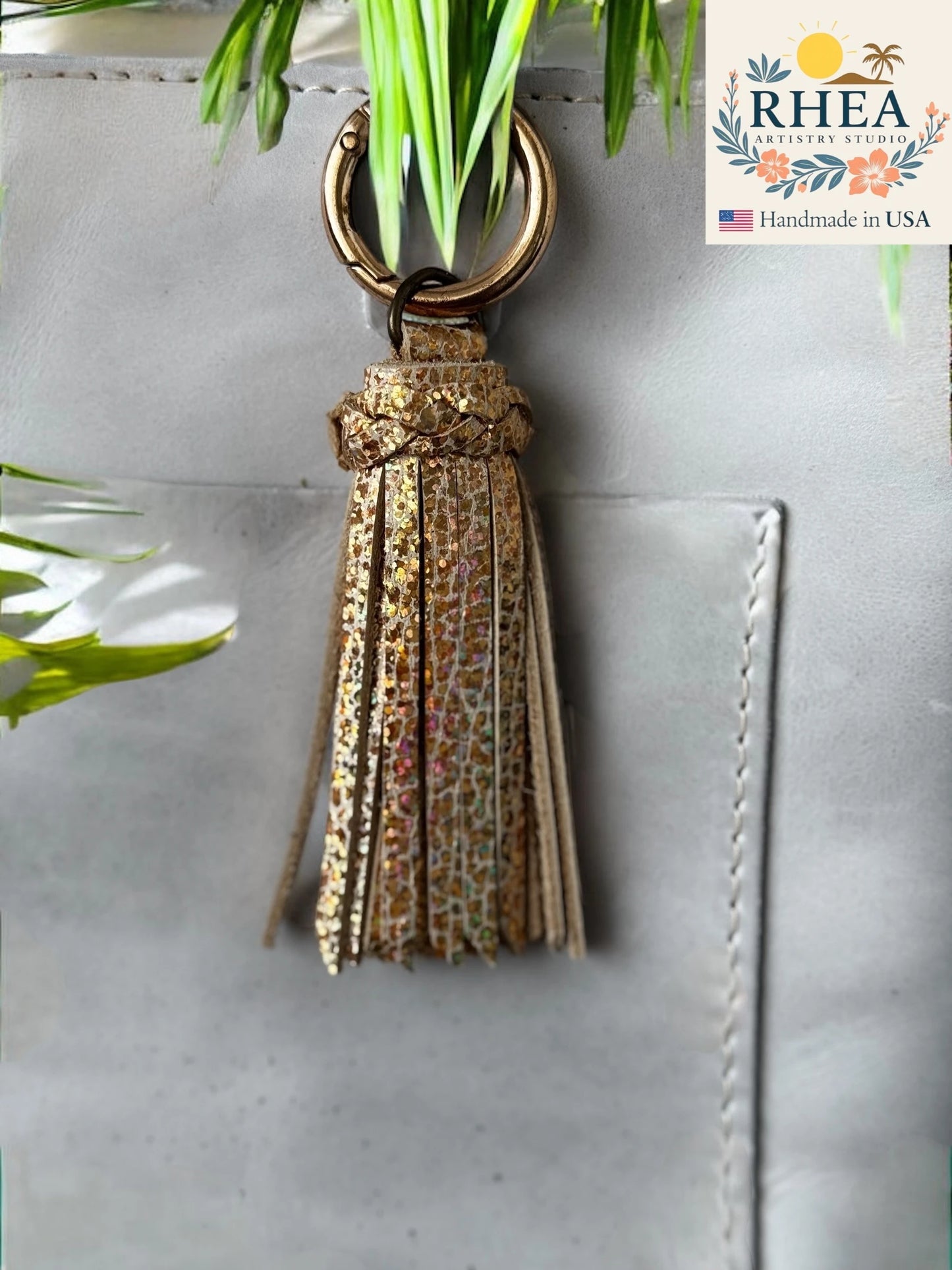 Gold tassel keychain on a gray surface with 'RHEA' branding.