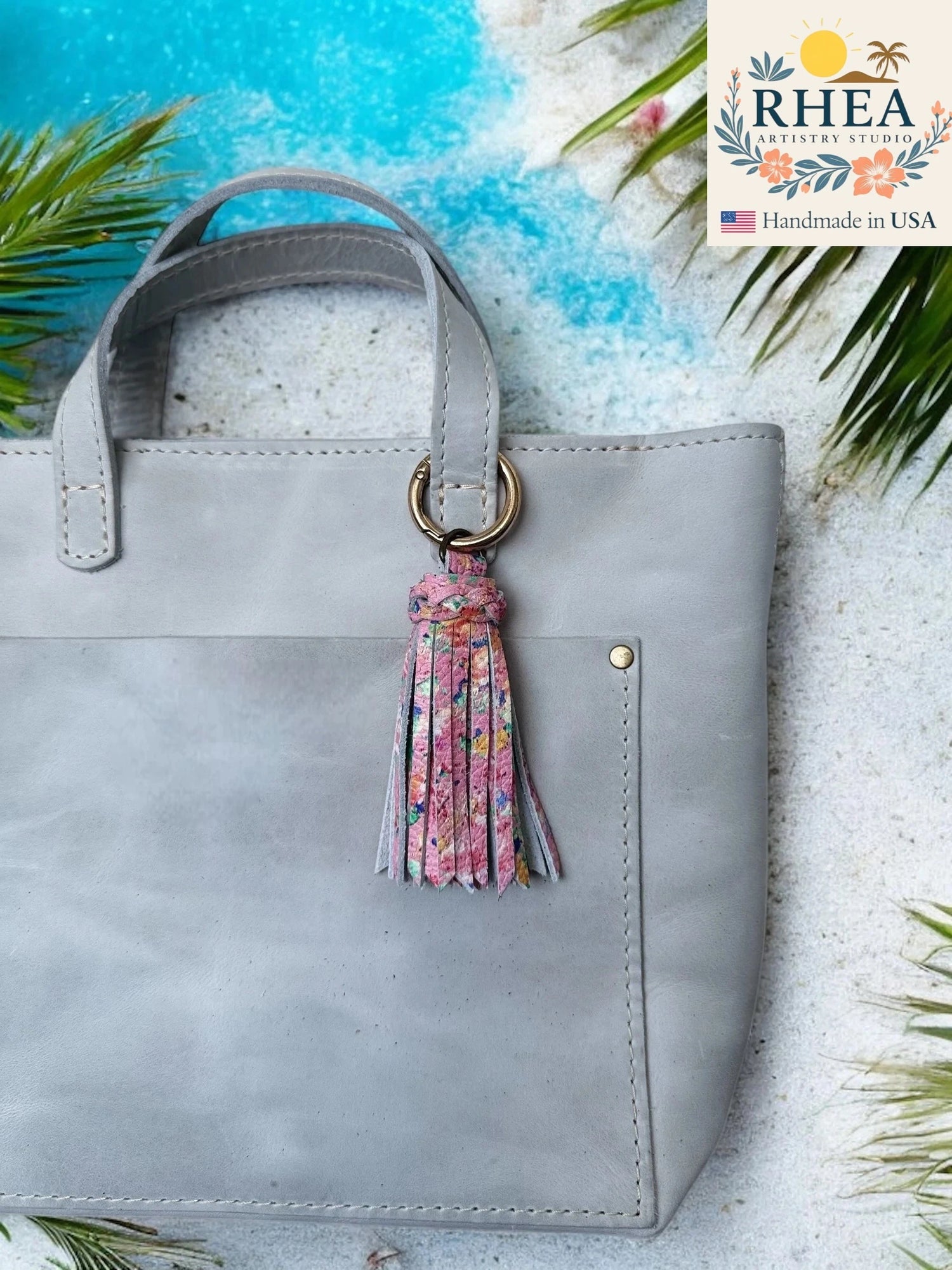 Gray handbag with a floral tassel on a textured surface with palm leaves and RHEA branding.