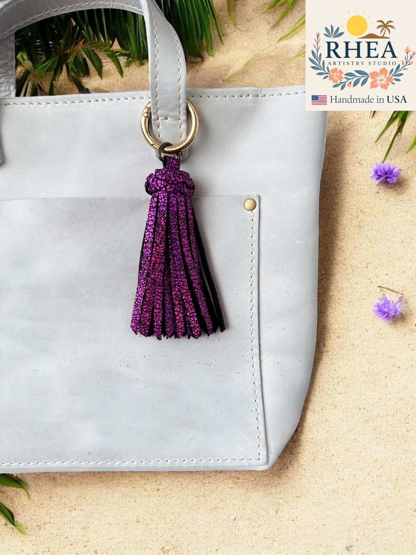 White handbag with a purple tassel keychain on a beige surface, RHEA brand visible.