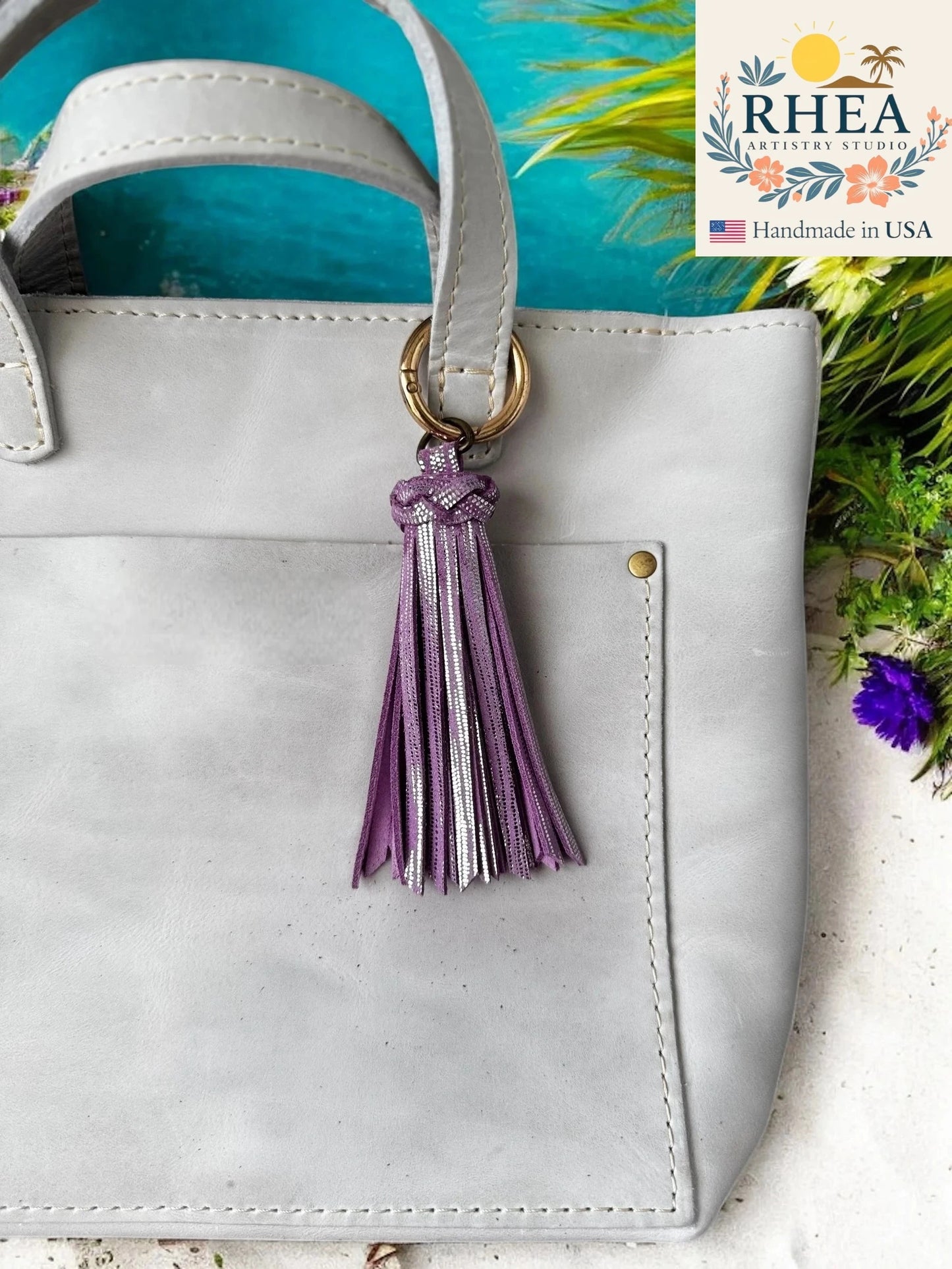 White handbag with a lilac tassel keychain on a blue background, featuring the RHEA brand.