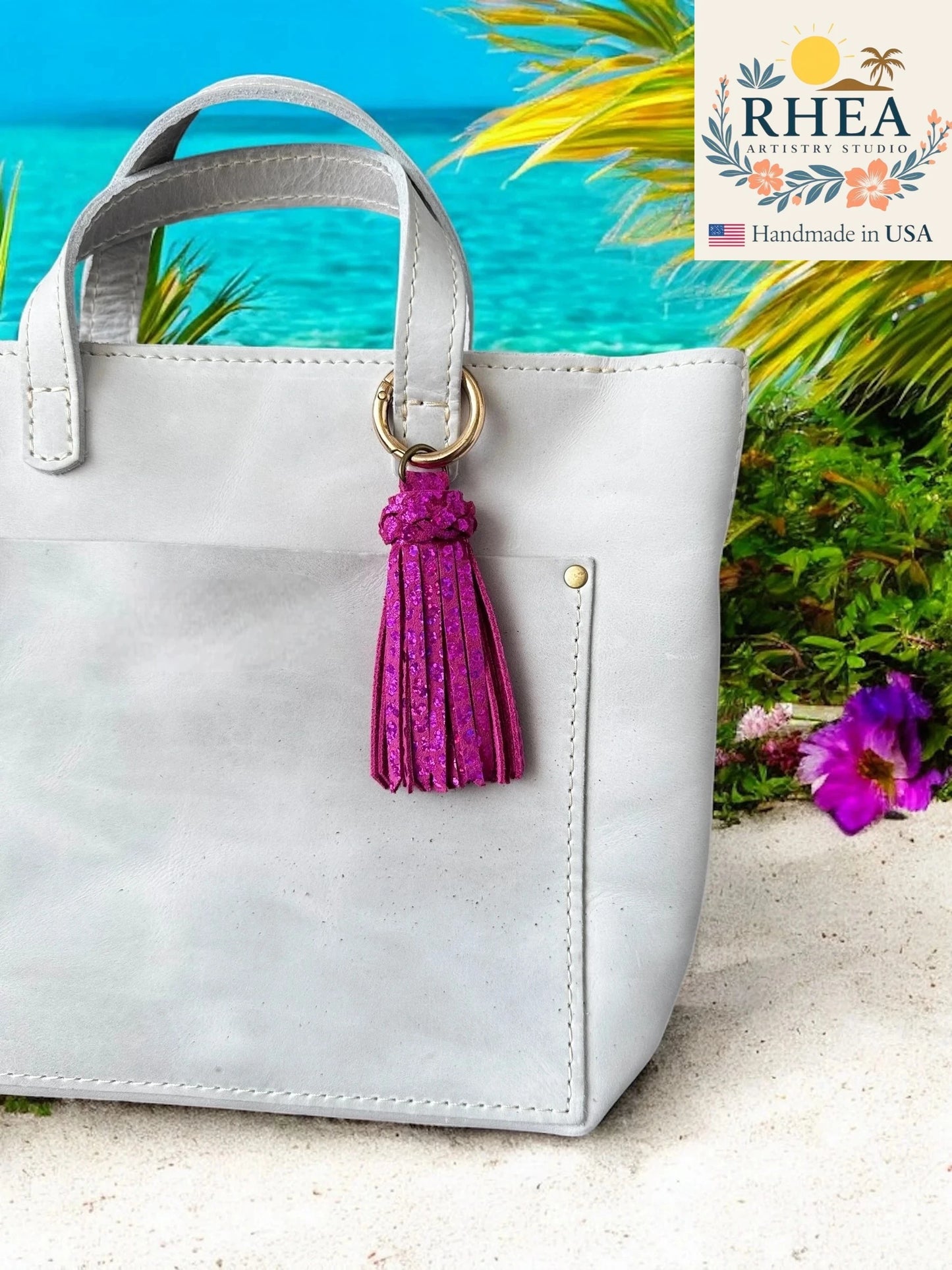 White handbag with a pink tassel on a beach setting, featuring the RHEA Artistry Studio logo.