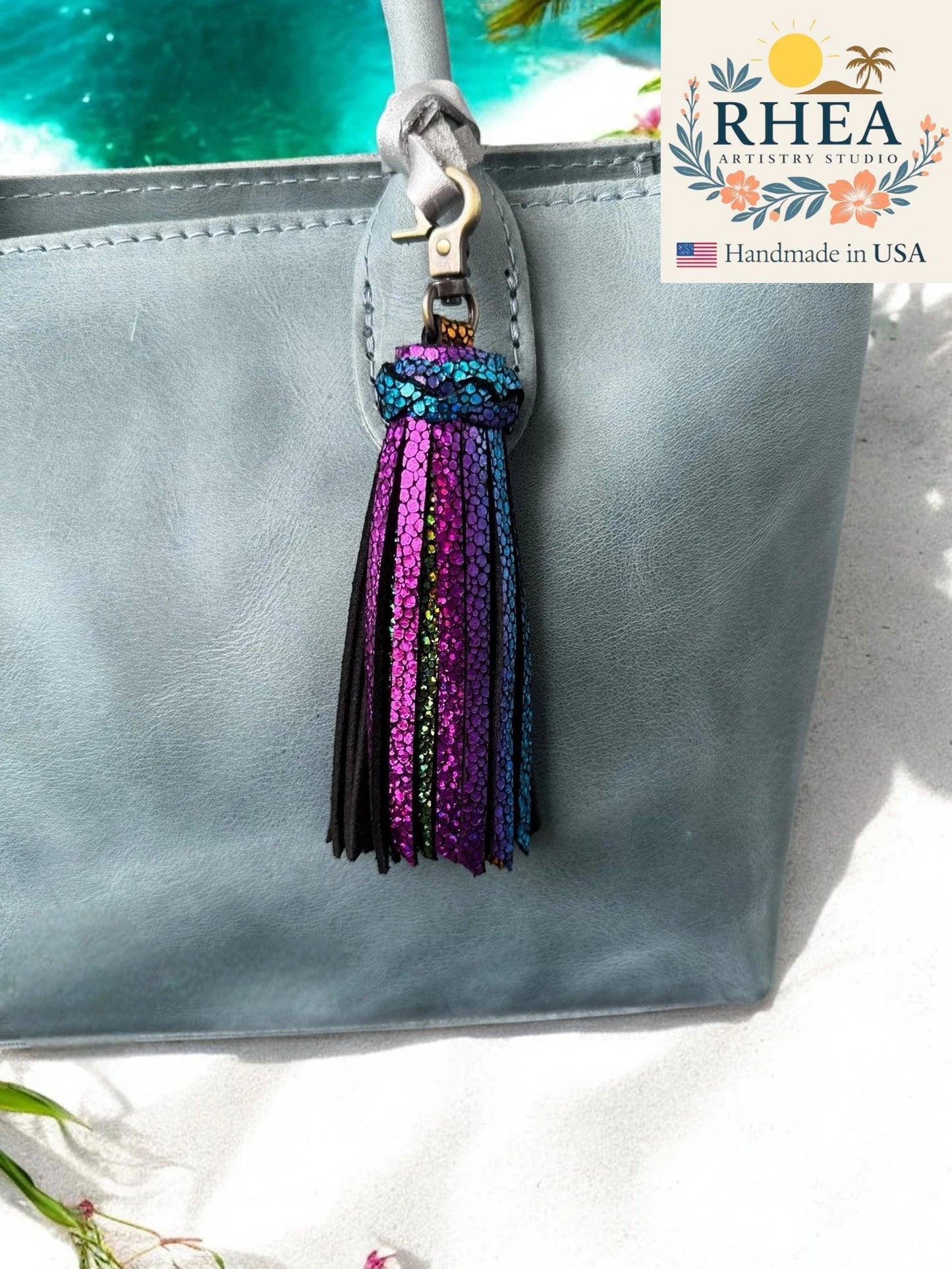 Gray bag with a colorful tassel keychain against a blurred natural background, featuring the RHEA Artistry Studio logo.