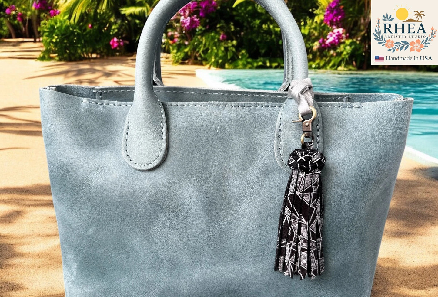Light blue handbag with a tassel keychain by RHEA, set against a poolside background.