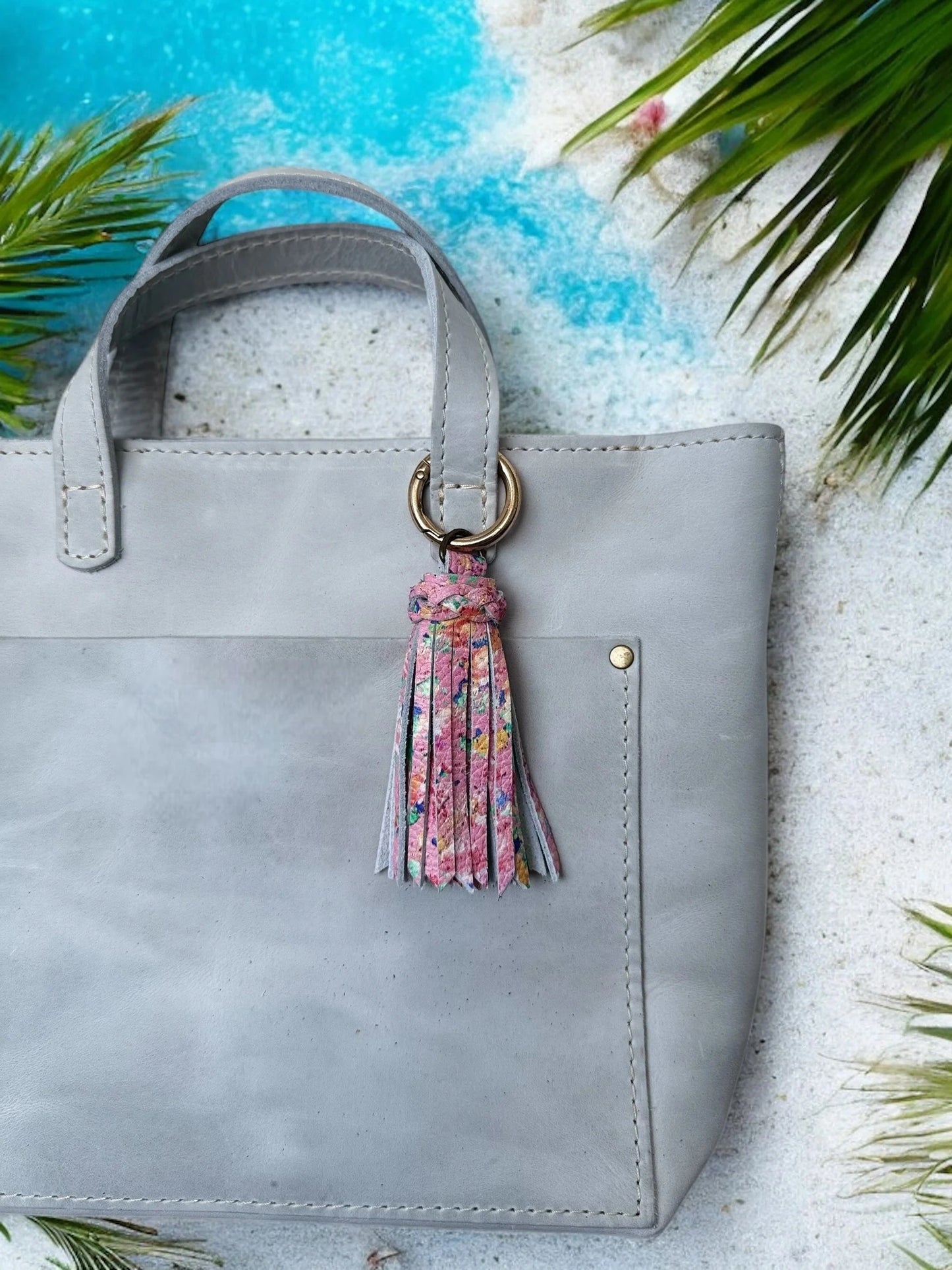 Gray handbag with a floral tassel on a sandy background with palm leaves
