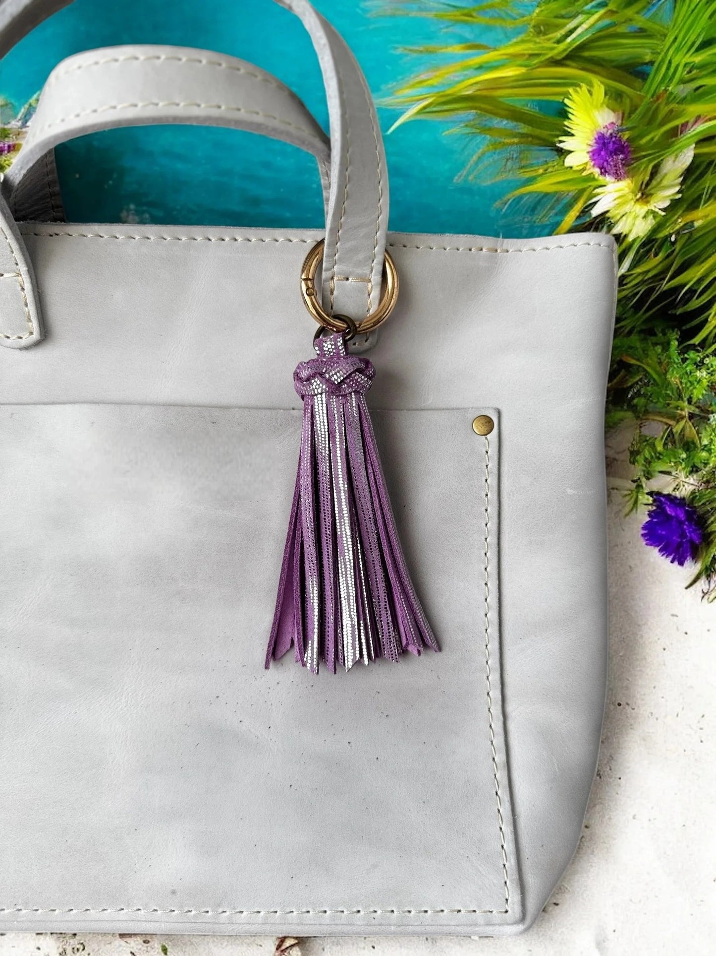 White handbag with a purple tassel keychain against a blue and green floral background