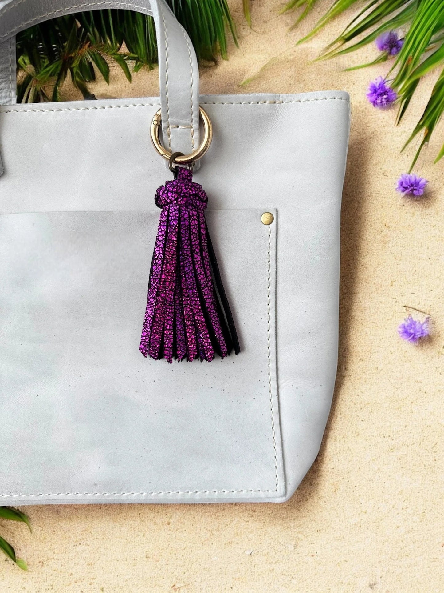 White handbag with a purple tassel keychain on a sandy surface with greenery.