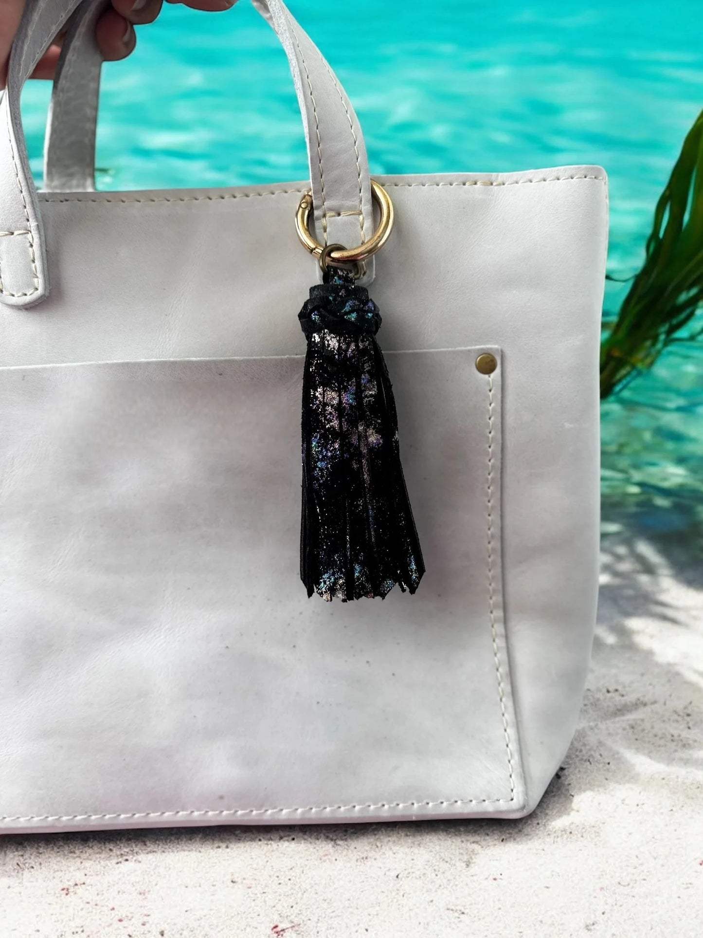 White handbag with a black tassel keychain on a sandy surface with water in the background