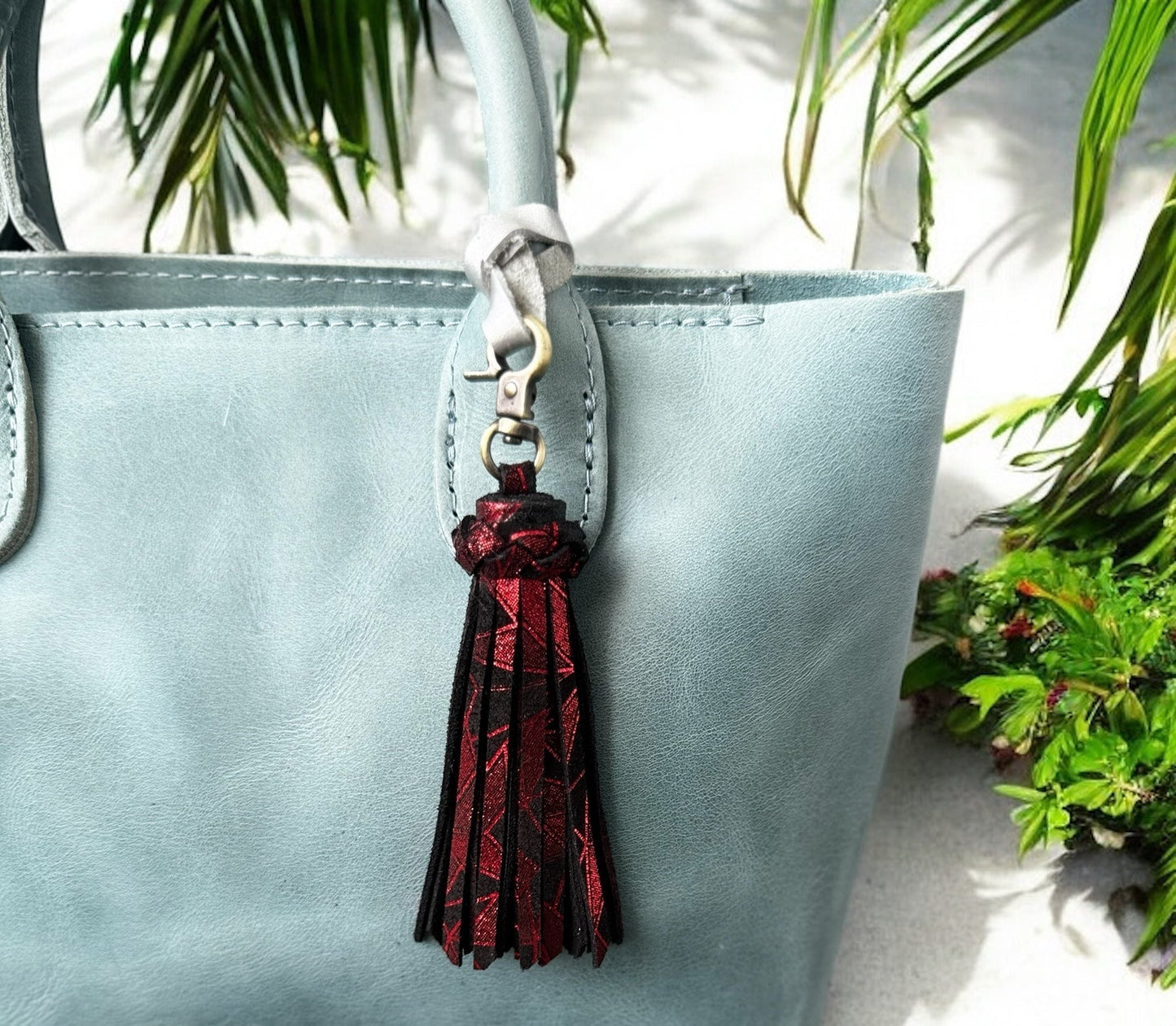 Light blue handbag with a red tassel keychain on a white surface with green plants in the background