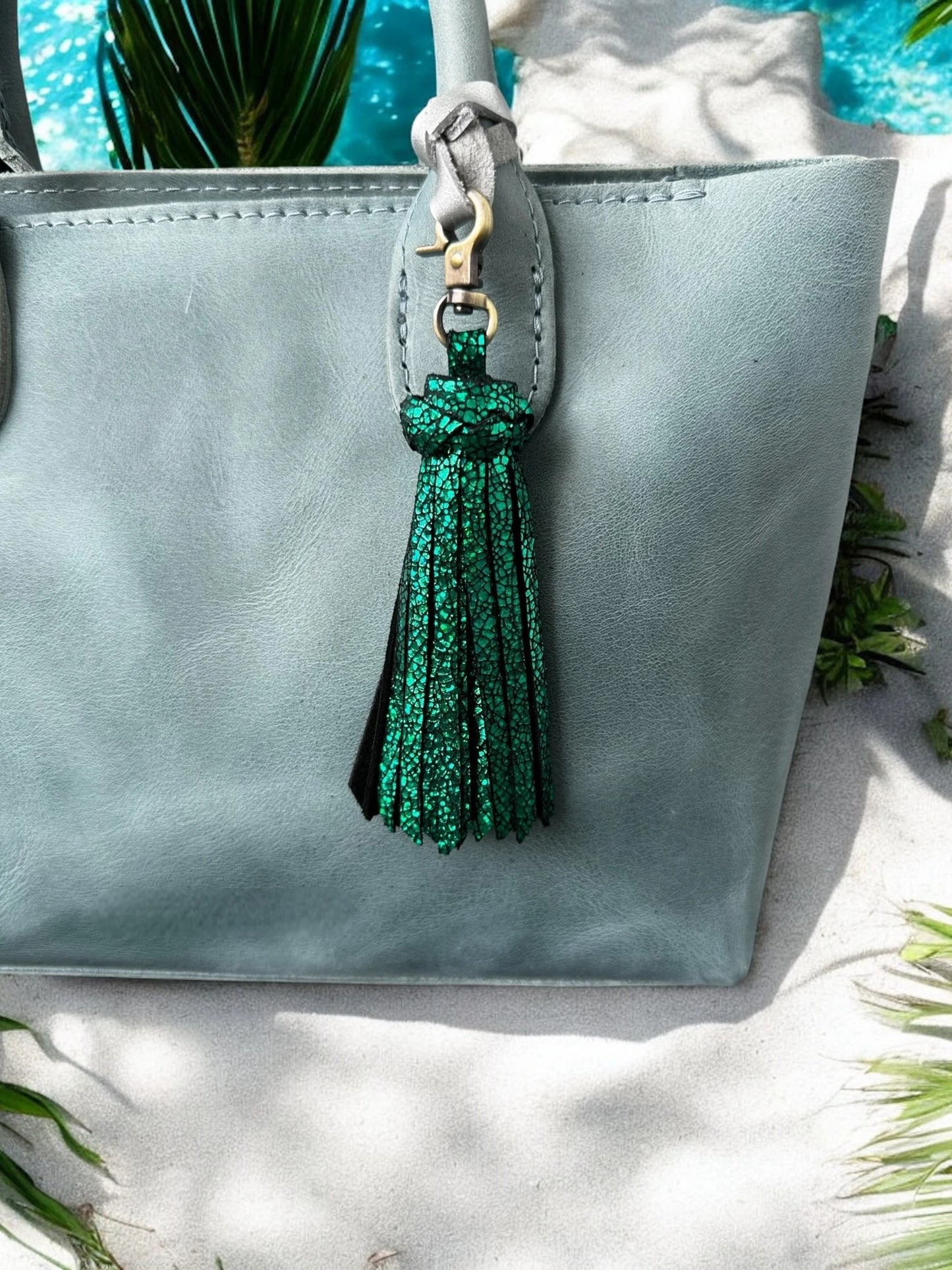 Light blue handbag with a green tassel on a white surface with greenery.