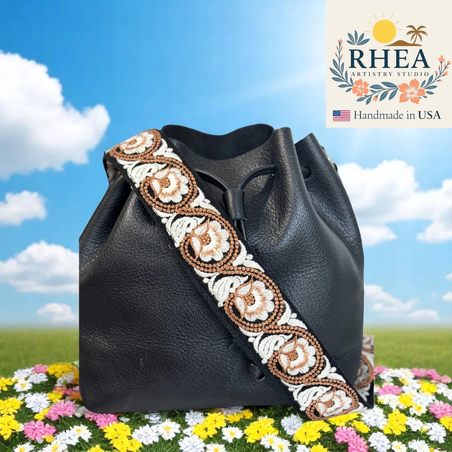 Black leather bag with decorative purse strap on a grassy field with flowers and blue sky.