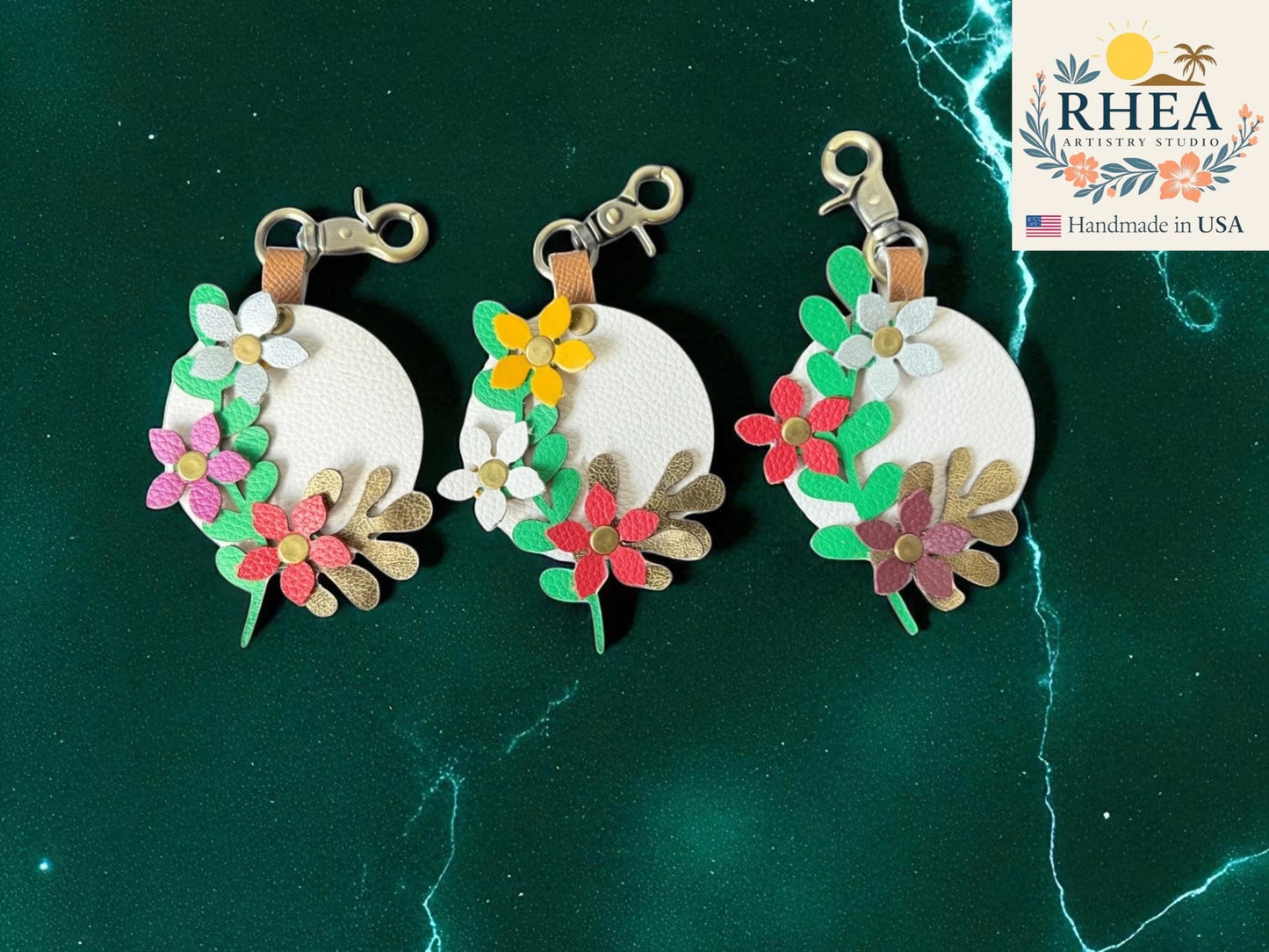 Three floral leather bag charms on a green marble background with RHEA branding.