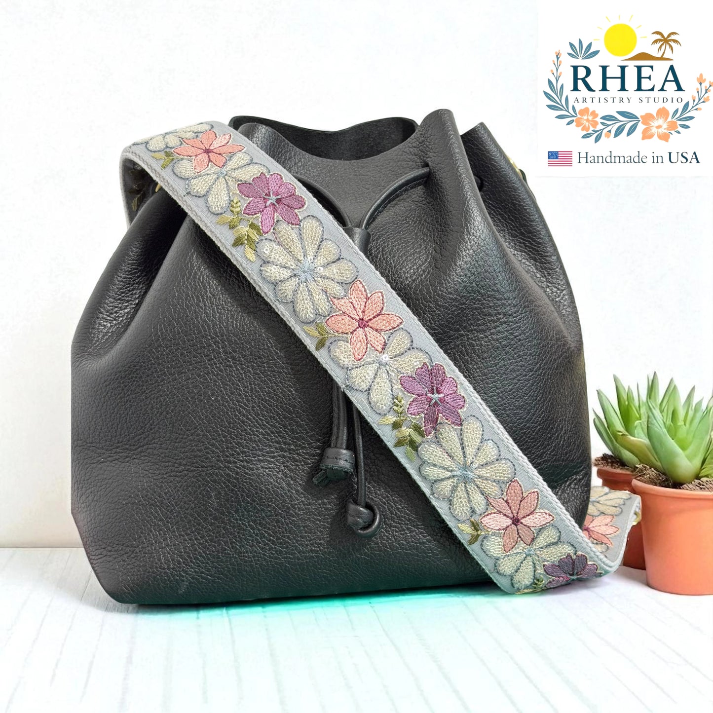 Black leather bag with a floral leather bag strap on a white background, featuring the RHEA brand.