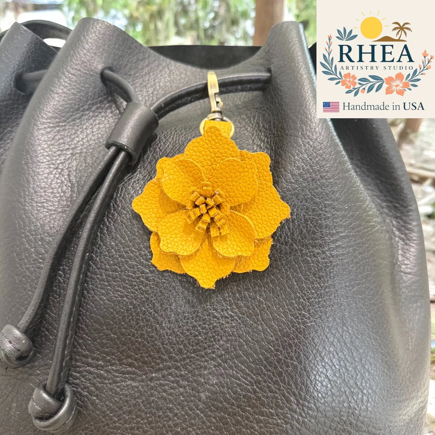 Mustard flower-shaped purse charm on a gray leather bag with RHEA Artistry Studio branding.