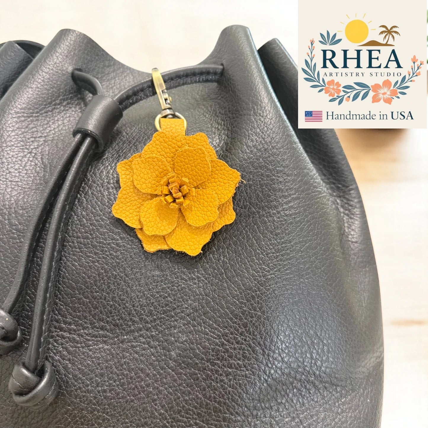 Mustard flower-shaped bag charm attached to a gray bag with RHEA Artistry Studio branding.