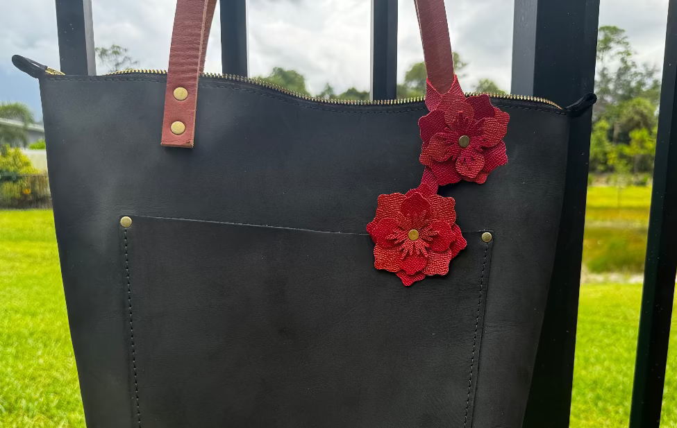 A red and brick red leather flower accessory with a textured surface and brass rivets, designed to attach to a bag or garment.