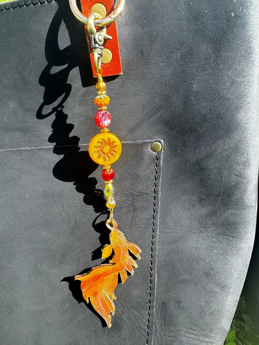 A handcrafted leather charm with a koi fish design, featuring solid brass hardware and high-end designer quality Czech glass, attached to a black leather strand.