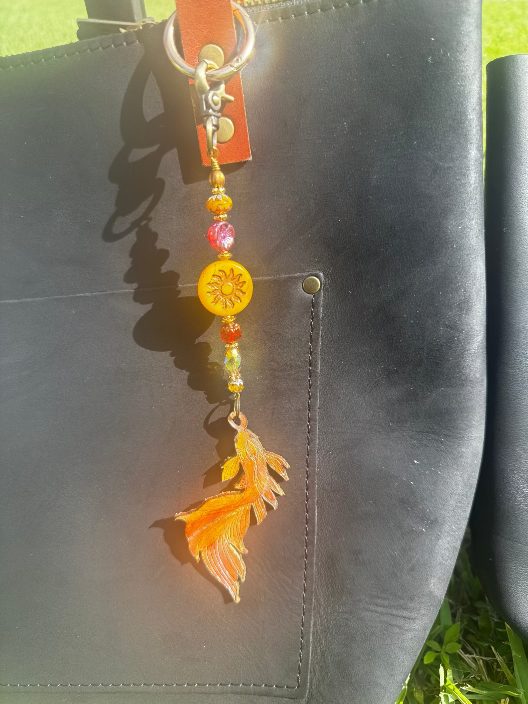 A handcrafted leather charm with a koi fish design, featuring solid brass hardware and high-end designer quality Czech glass, attached to a black leather strand.