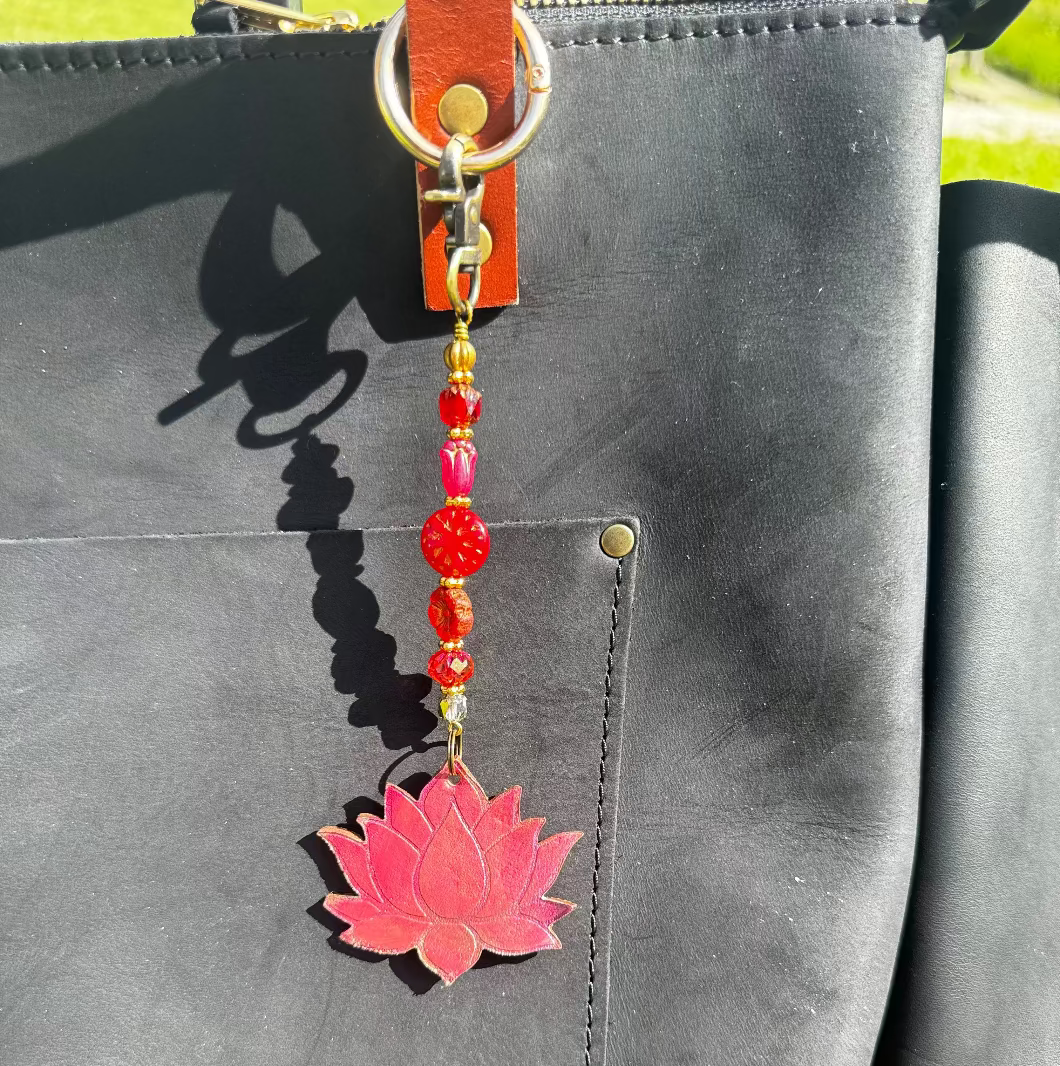 A handcrafted lotus bloom charm made of red and orange glass with brass hardware, attached to a leather-bound notebook.