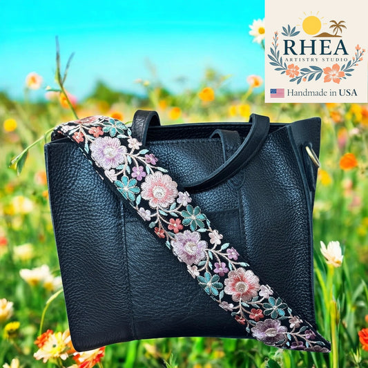 Black handbag with floral purse strap in a field of flowers, featuring the RHEA Artisan Studio logo.