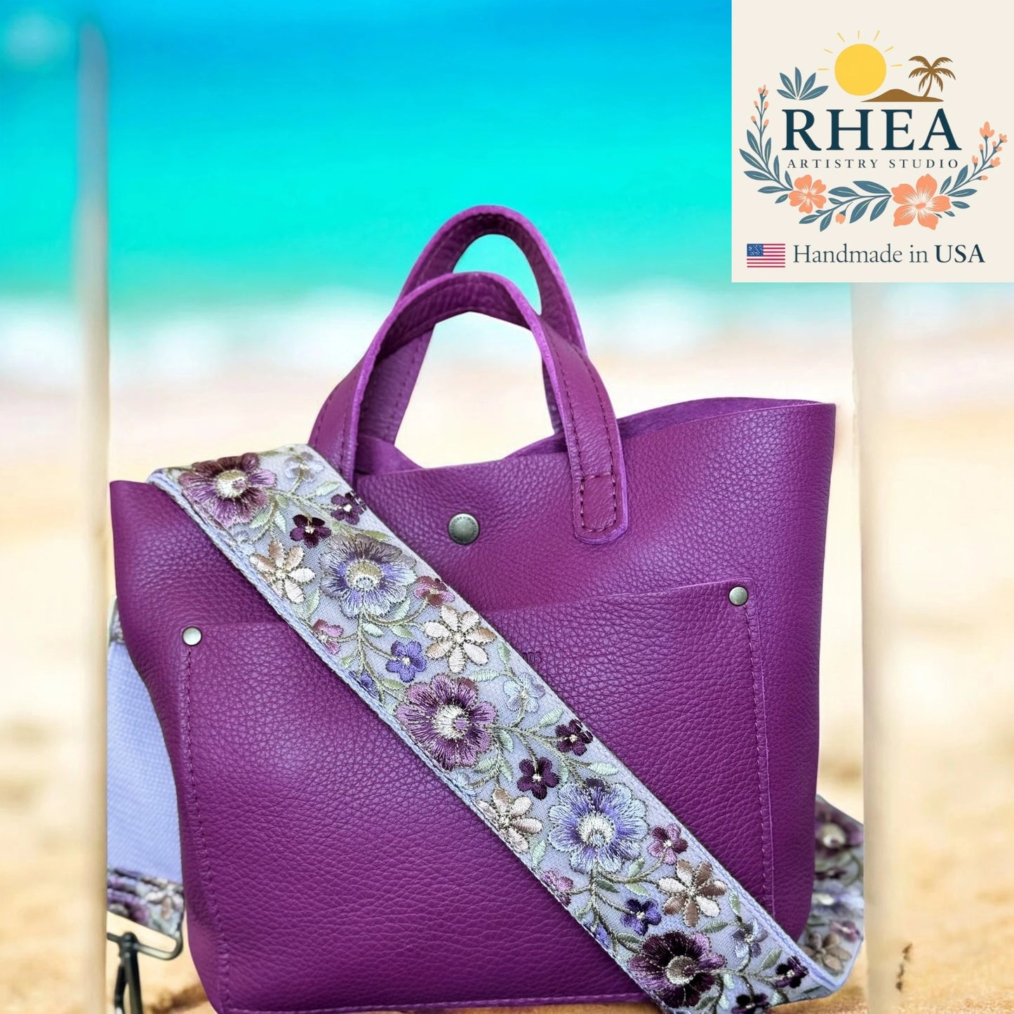 Purple handbag with a floral bag strap on a beach background, featuring the RHEA Artisan Studio logo.